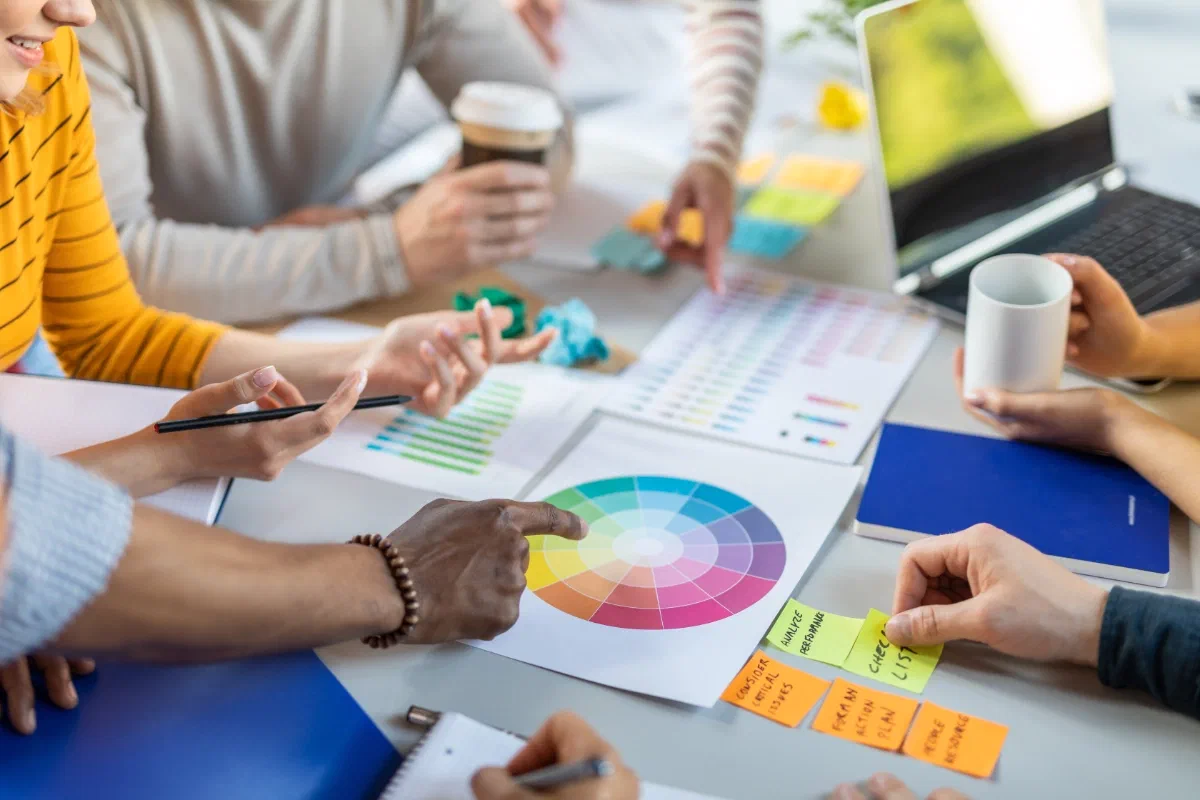 A team around a desk covered in colour wheels, printed colour charts and sticky notes, one hand pointing at a colour wheel