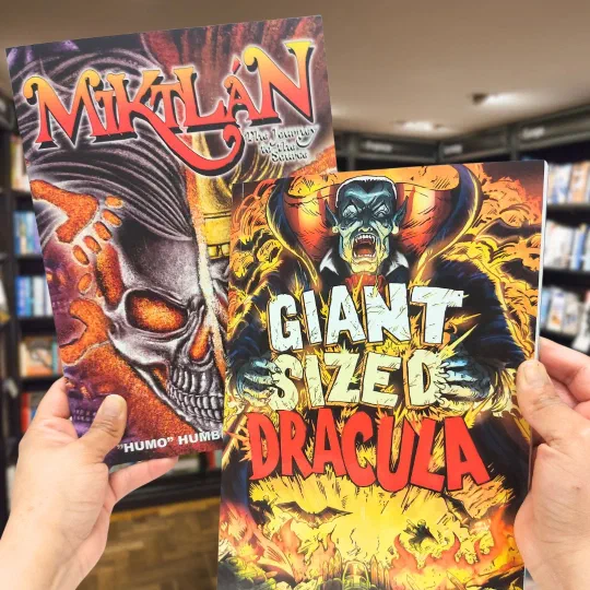 Two graphic novels held up in a bookshop, Miktán and Giant Sized Dracula